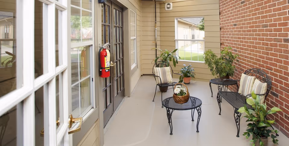 A covered outdoor patio area with a black metal table, two black metal chairs with striped cushions, a black metal bench with striped cushions, and several potted plants. The patio is adjacent to a brick wall and beige siding with windows and a door that has a fire extinguisher mounted beside it.