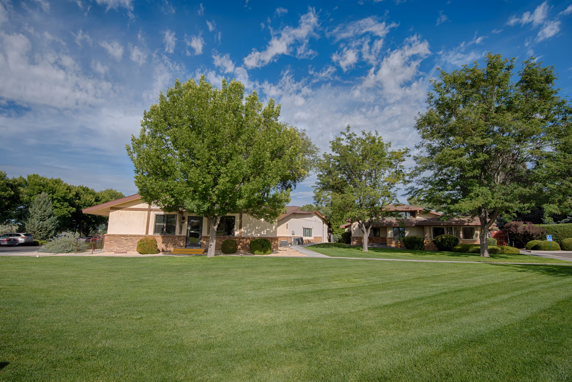 A wide view of a senior living facility's exterior with two single-story buildings surrounded by green grass and several trees under a partly cloudy blue sky.