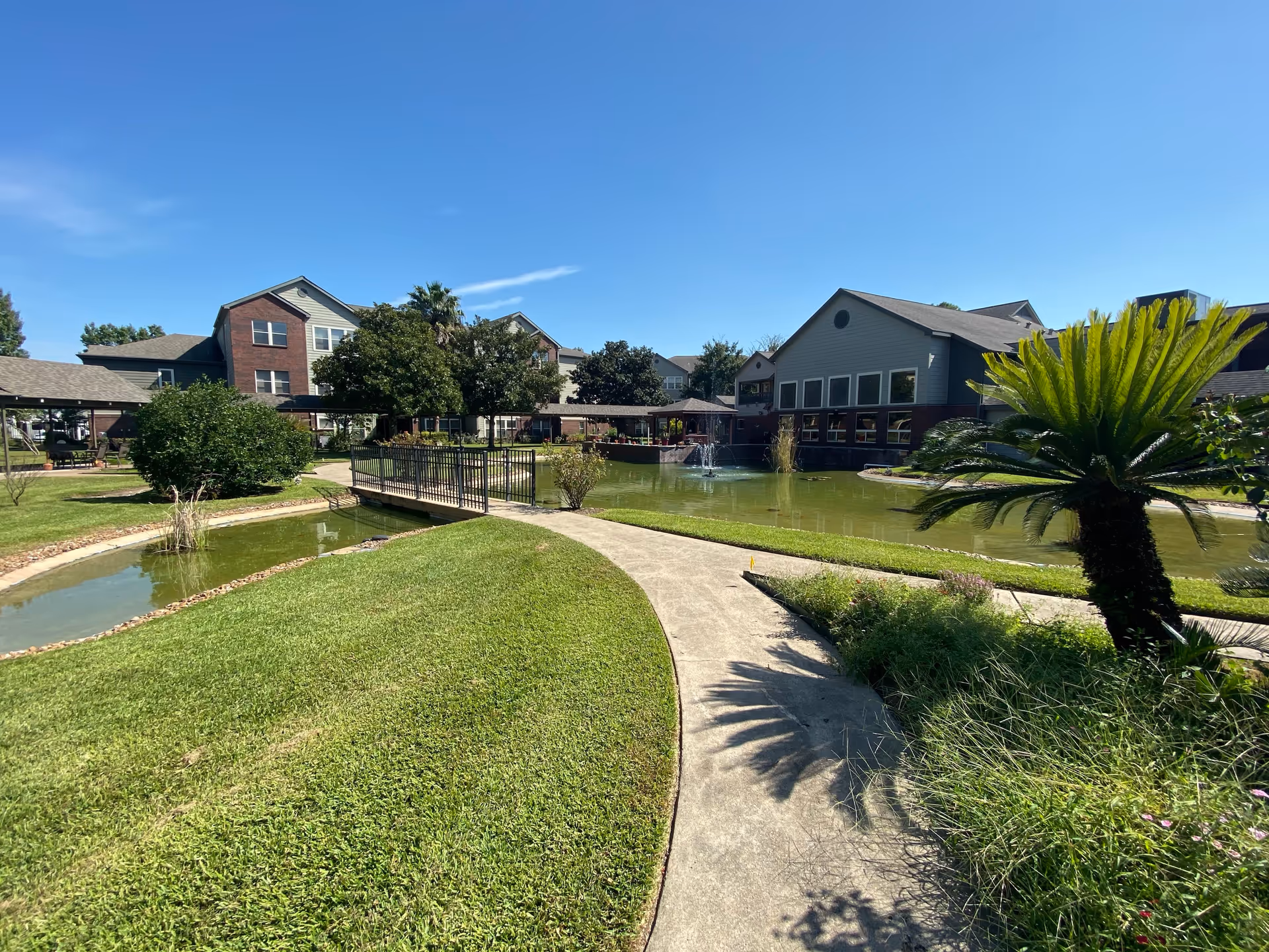 Curved walkway and small bridge over a pond with a fountain in front of multi-story senior living buildings and landscaping under a clear blue sky.