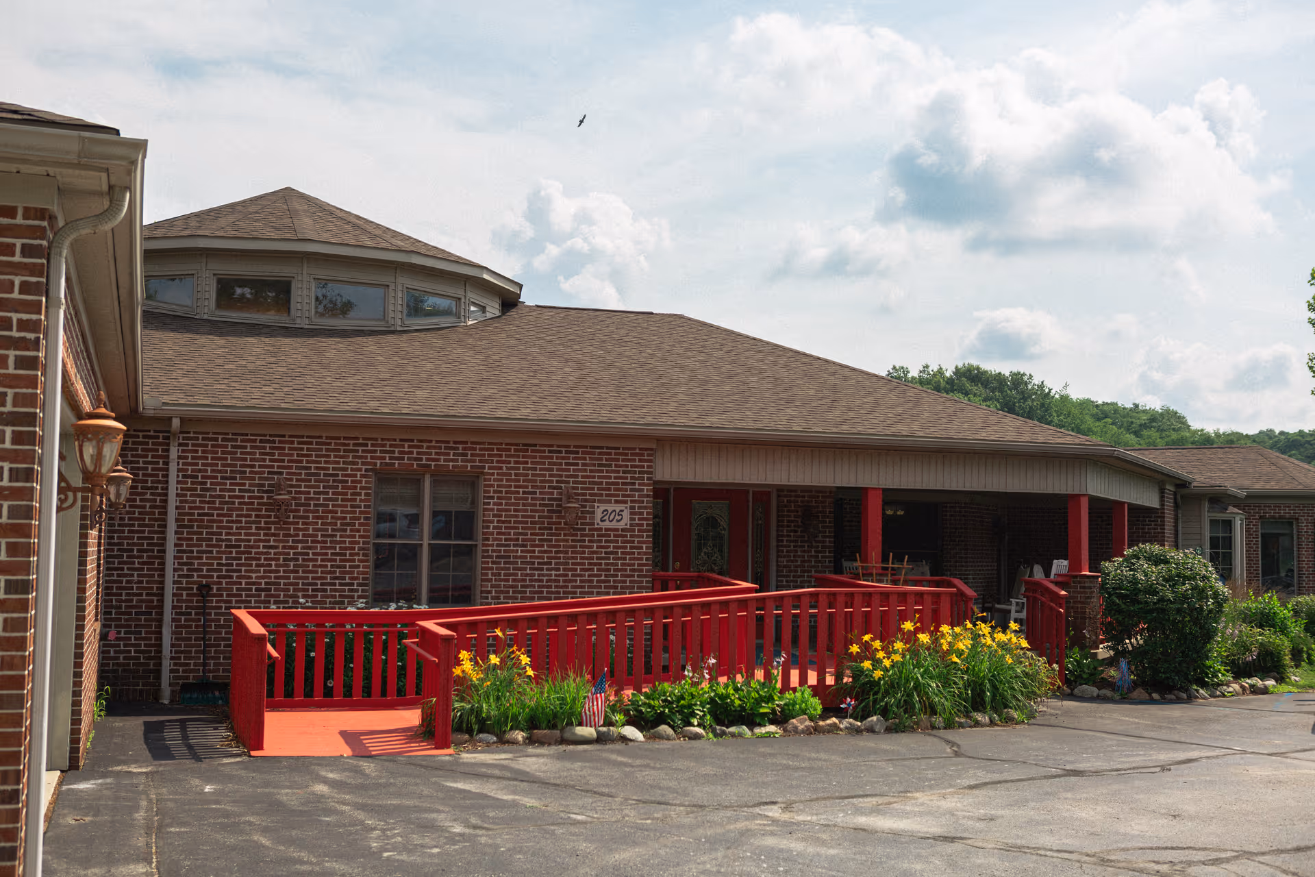 Exterior view of a brick building with a red wooden ramp and railing leading to a covered porch area. The building has a brown shingled roof and a small garden with yellow flowers and greenery in front. The sky is partly cloudy.