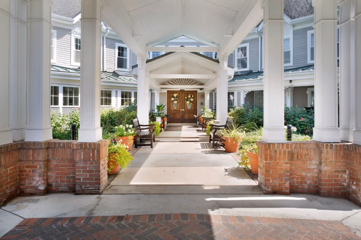Covered entrance walkway leading to double wooden doors decorated with wreaths, flanked by benches and potted plants, with a building featuring multiple windows and a green metal roof in the background.