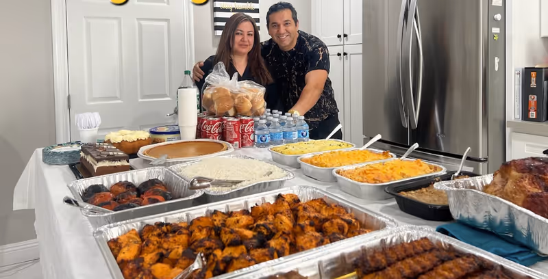 Two people stand behind a long buffet table laden with trays of prepared foods inside a kitchen.