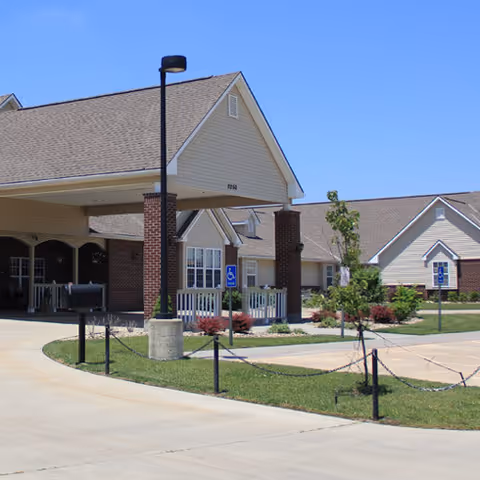 Exterior view of a senior living facility with beige siding and brick columns, a covered entrance with a driveway, handicap parking signs, small trees, and landscaping under a clear blue sky.