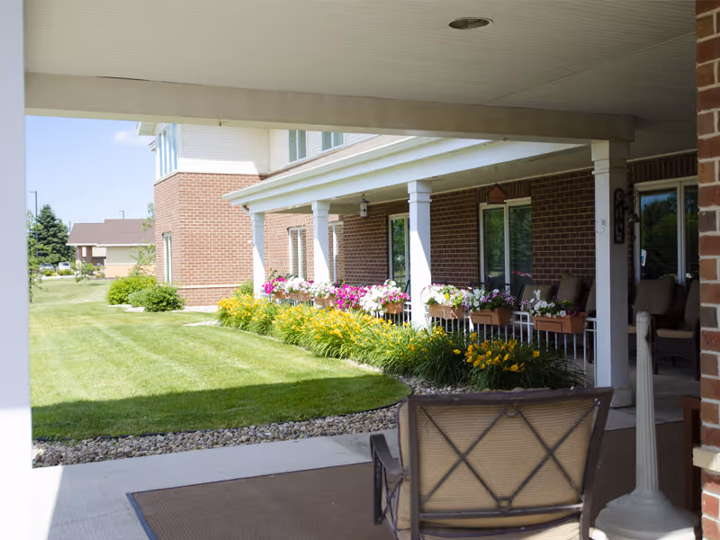 View of a covered outdoor patio area with chairs and flower boxes filled with colorful flowers along the edge. The patio is attached to a brick building with white columns supporting the roof. There is a well-maintained lawn and garden with yellow flowers adjacent to the patio.