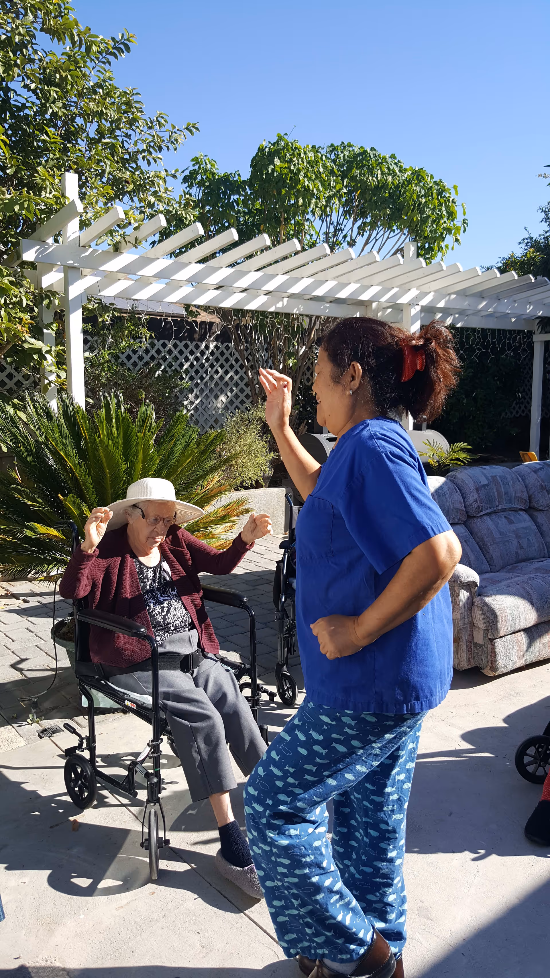 An elderly woman sitting in a wheelchair wearing a white hat and a maroon cardigan, raising her arms outdoors under a white pergola. A caregiver in blue scrubs and patterned pants is standing nearby, engaging with the elderly woman. There are green plants and a couch in the background under a clear blue sky.