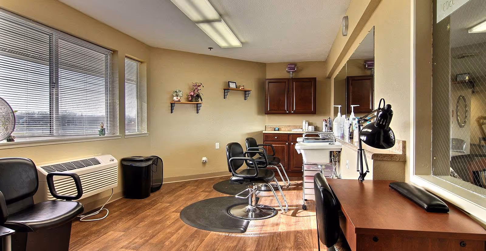 Interior view of a hair salon area in a senior living facility with three black salon chairs, a hair washing station, a wooden desk with a black lamp, and large windows with blinds letting in natural light. The room has wooden flooring, beige walls, and some small decorative shelves with plants and flowers.