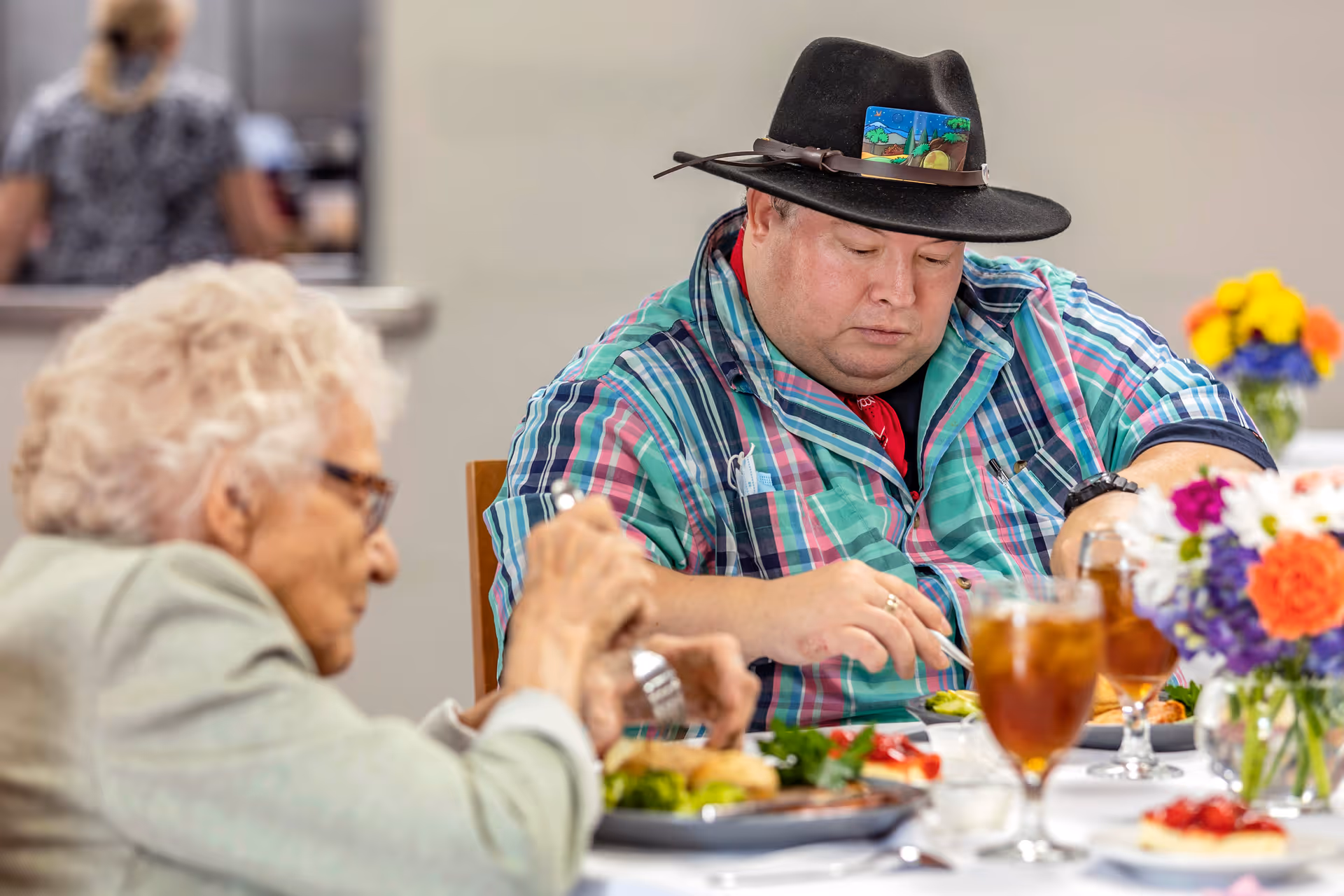 Two people sitting at a dining table enjoying a meal together. One person is an elderly woman with white hair and glasses, and the other is a man wearing a black hat with a colorful band and a plaid shirt. The table is set with plates of food, glasses of iced tea, and a vase with colorful flowers.