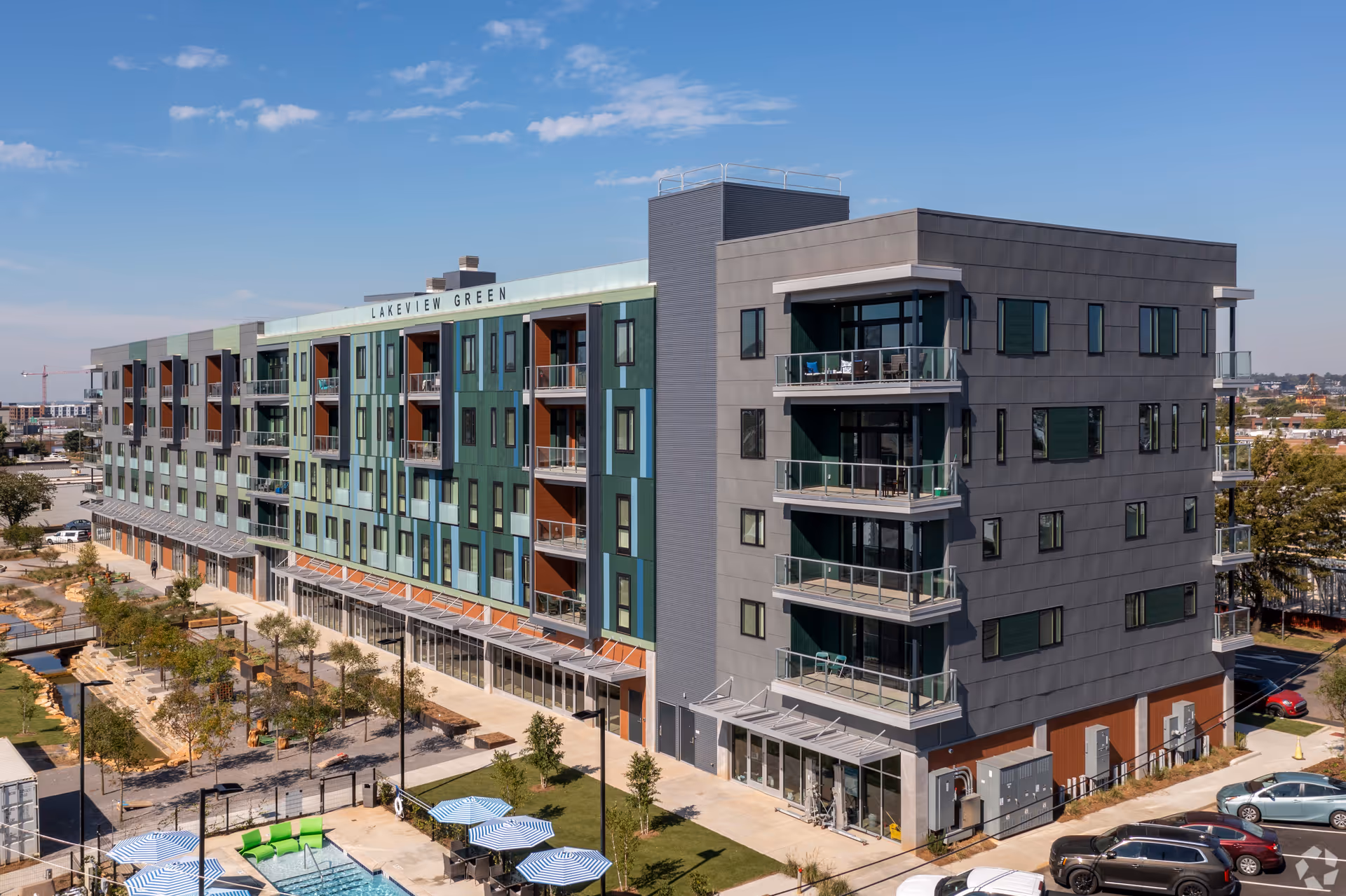 Exterior view of a modern multi-story residential building named Lakeview Green with balconies, large windows, and a landscaped area including a pool with blue and white umbrellas and green seating. Several cars are parked in the adjacent parking lot under a clear blue sky.