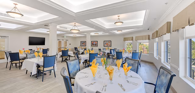 A bright and spacious dining room with multiple round tables covered in white tablecloths. Each table is set with yellow folded napkins, glassware, and silverware. Blue cushioned chairs surround the tables. Large windows on one side allow natural light to fill the room. The ceiling features recessed lighting and decorative light fixtures. There is a flat-screen TV mounted on one wall and framed artwork hanging on another.