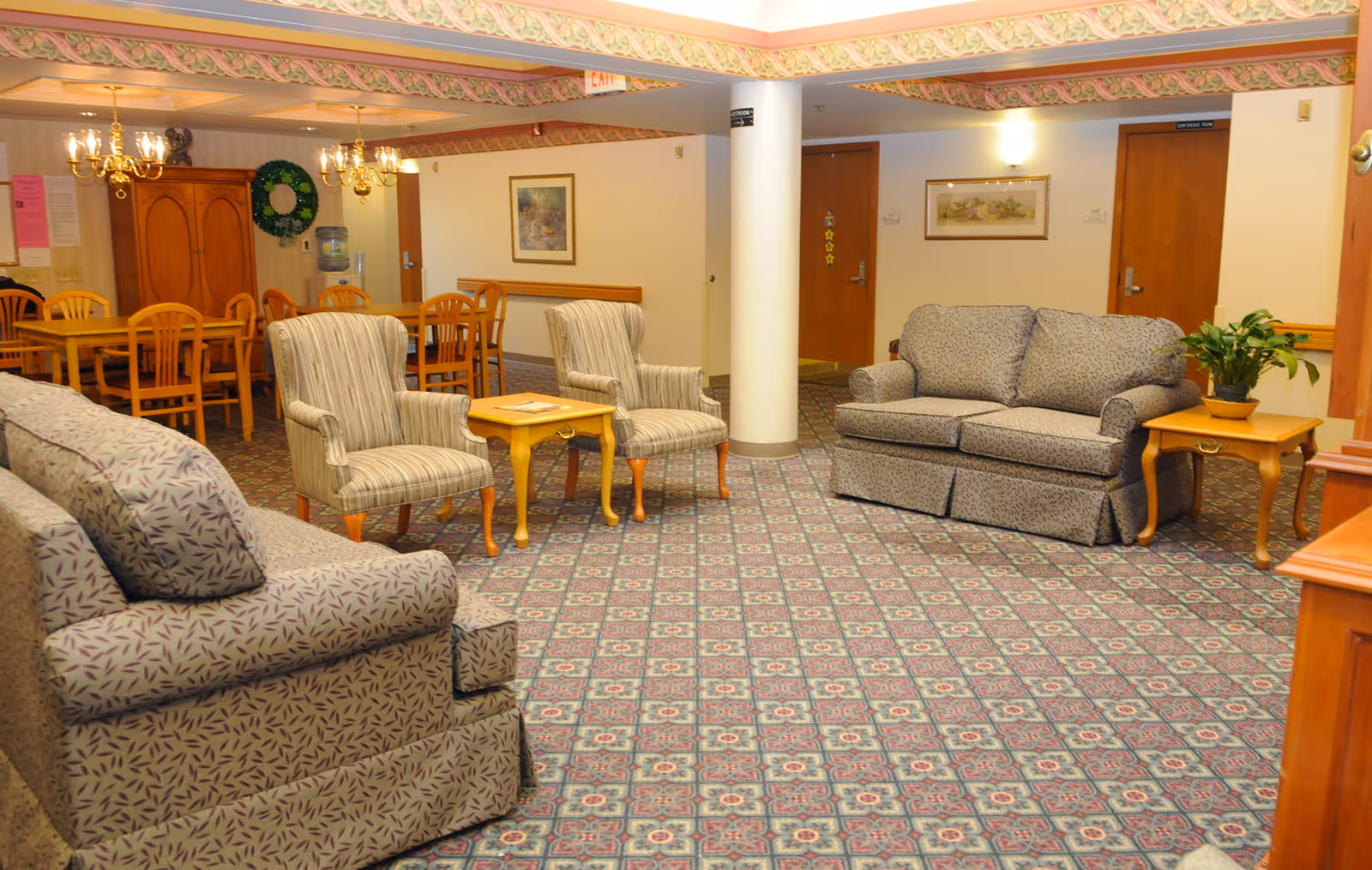 A cozy common area in a senior living facility featuring two patterned sofas, two striped armchairs, and two wooden side tables with a plant and a book. In the background, there is a dining area with wooden tables and chairs, chandeliers, a wooden cabinet, and framed artwork on the walls.