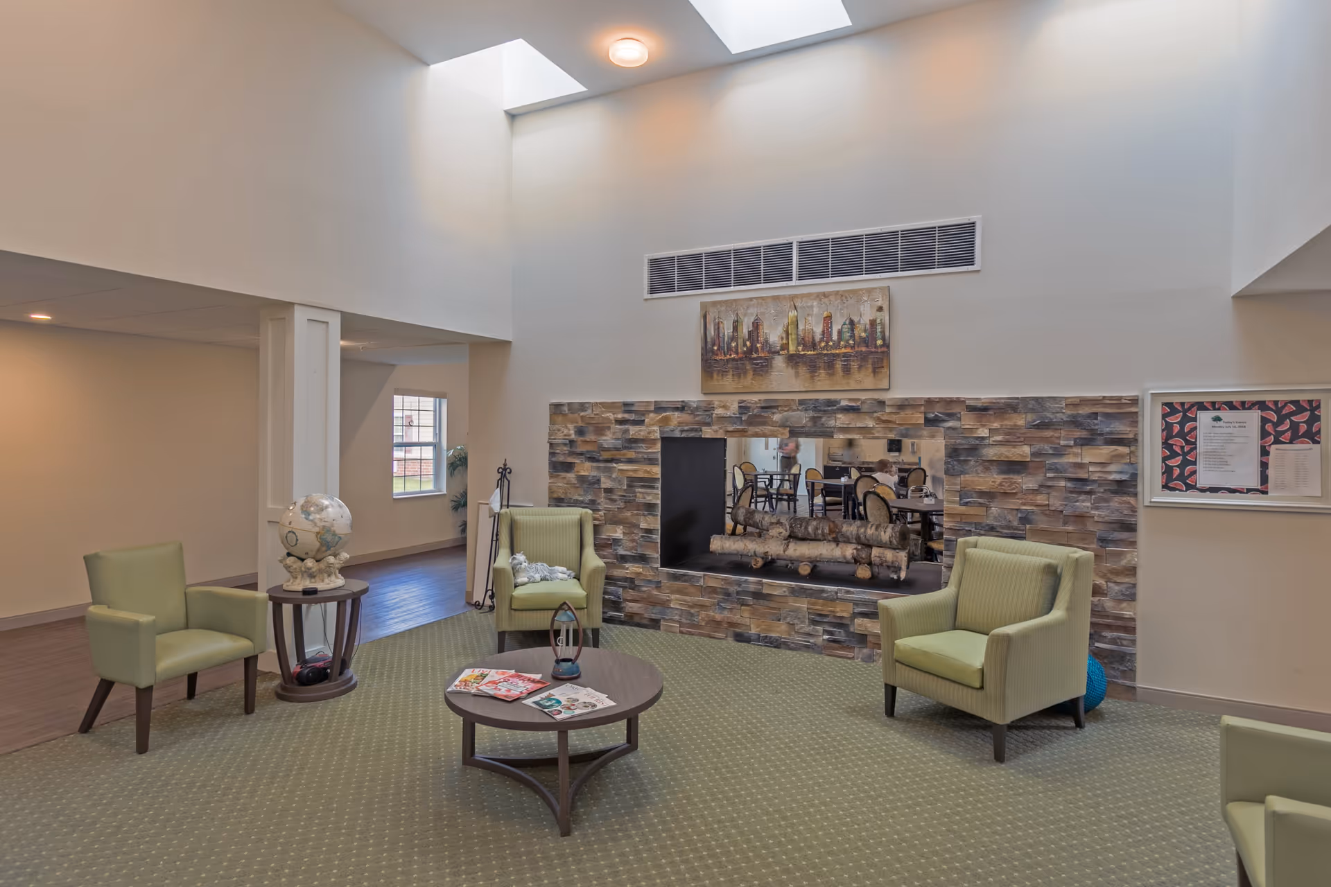 A cozy living room area in a senior living facility with green upholstered armchairs arranged around a circular wooden coffee table holding magazines and a decorative lantern. The room features a stone accent wall with a built-in fireplace and a cityscape painting above it. There is a skylight allowing natural light to brighten the space, and a globe on a small side table near one of the chairs.
