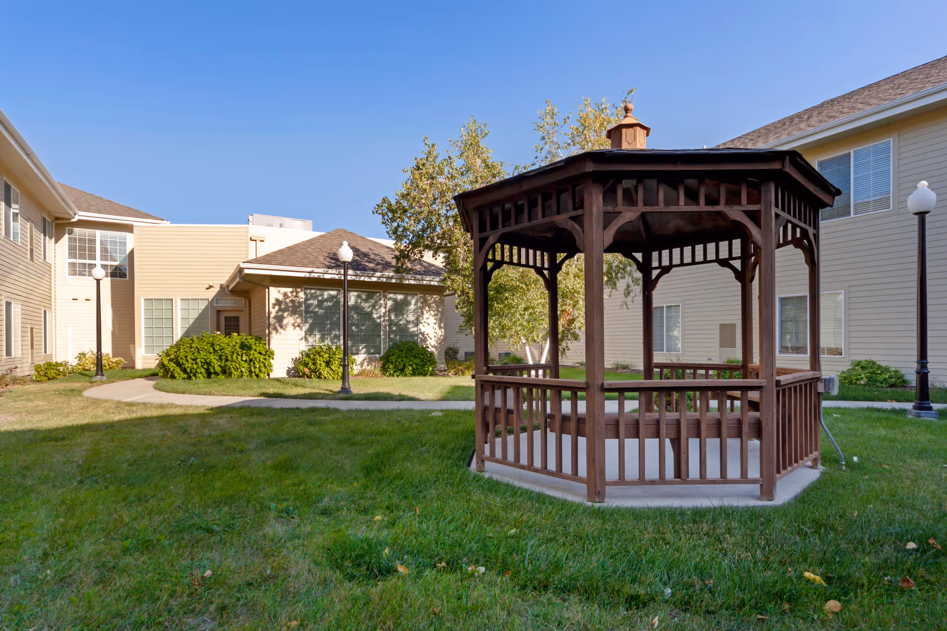Outdoor courtyard area of Elk Ridge Village Assisted Living featuring a wooden gazebo on a concrete base surrounded by green grass, with beige two-story buildings and lamp posts in the background under a clear blue sky.