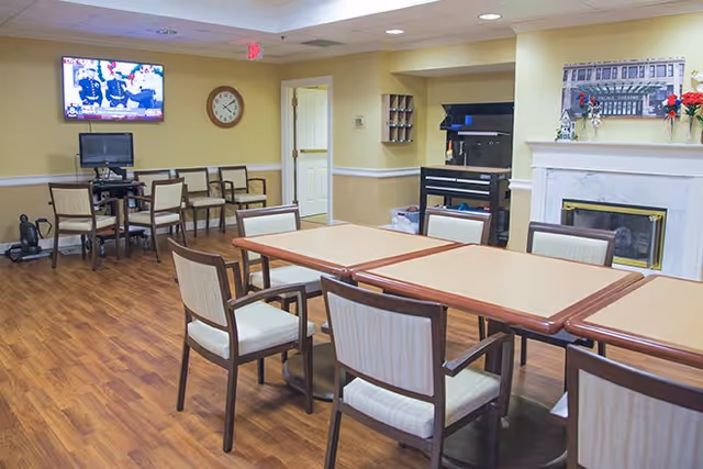 A common area in Richmond Heights Place featuring a rectangular table with several chairs around it, a fireplace with a mantel decorated with flowers and a framed picture above it, a wall-mounted TV showing a program, a clock on the wall, and additional chairs and a computer station in the background. The room has wood flooring and soft yellow walls.
