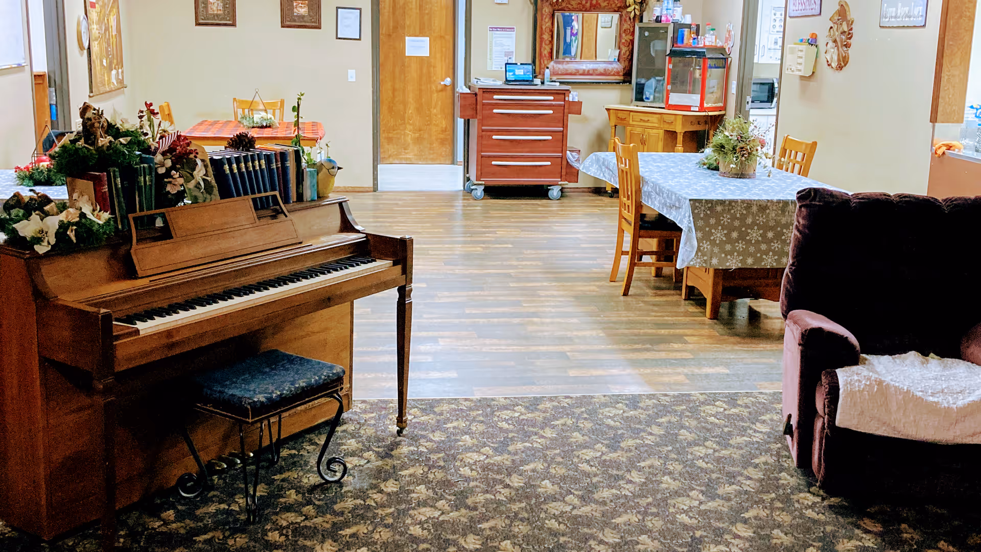 Interior of a senior living facility room featuring a wooden piano with a cushioned bench, decorated with plants and books. In the background, there is a dining table covered with a patterned tablecloth and surrounded by wooden chairs. To the right, there is a plush armchair with a white throw. The floor is a combination of patterned carpet and wood laminate. The walls have framed pictures and a wooden door is visible at the back.