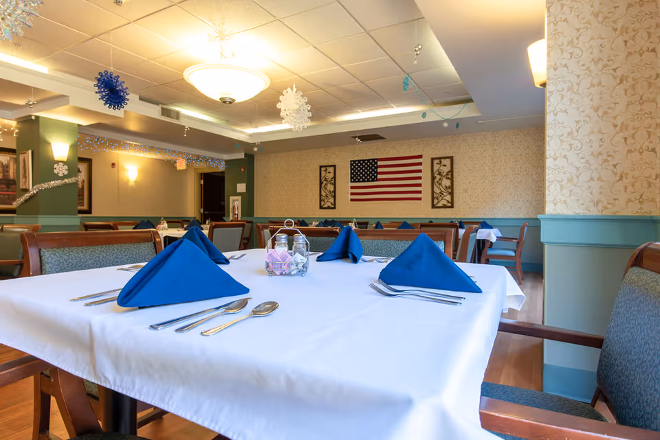 A dining room with tables covered in white tablecloths and set with blue folded napkins, silverware, and salt and pepper shakers. The room has wooden chairs with patterned upholstery, beige wallpaper with a floral design, and an American flag hanging on the far wall. Ceiling lights and hanging snowflake decorations are visible.