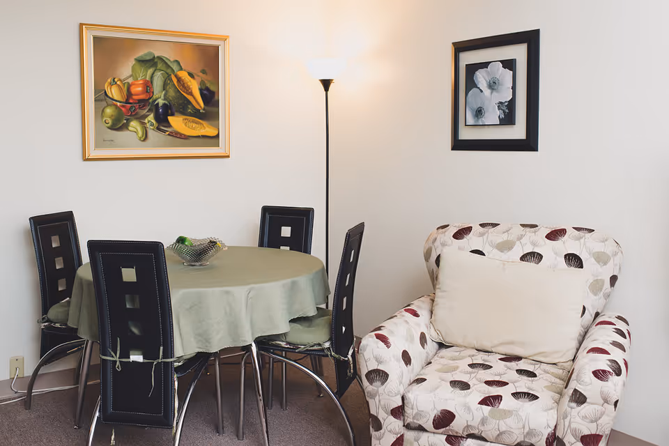 A cozy corner of a senior living facility room featuring a round dining table covered with a green tablecloth and surrounded by four black chairs. Next to the table is a patterned armchair with a large beige cushion. The walls are decorated with two framed pictures: one colorful painting of fruits and vegetables, and one black and white floral photograph. A tall floor lamp stands between the table and the armchair, providing soft lighting.