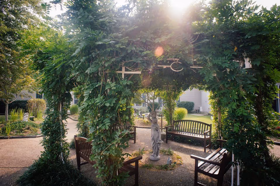 A peaceful outdoor garden area with a leafy green archway surrounding a small statue in the center. There are wooden benches arranged around the statue on a paved pathway, with trees and shrubs in the background under soft sunlight.