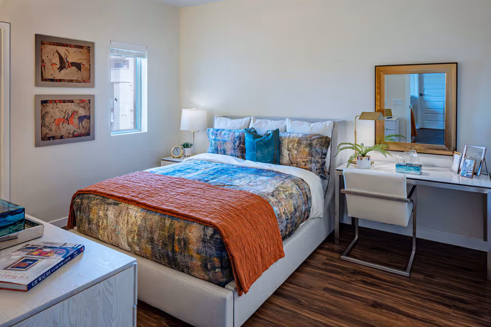 A neatly made bed with colorful bedding and an orange throw blanket in a bedroom. The room features a wooden floor, a white desk with a chair, a gold-framed mirror, a table lamp, and framed artwork on the wall. A small window allows natural light into the room.