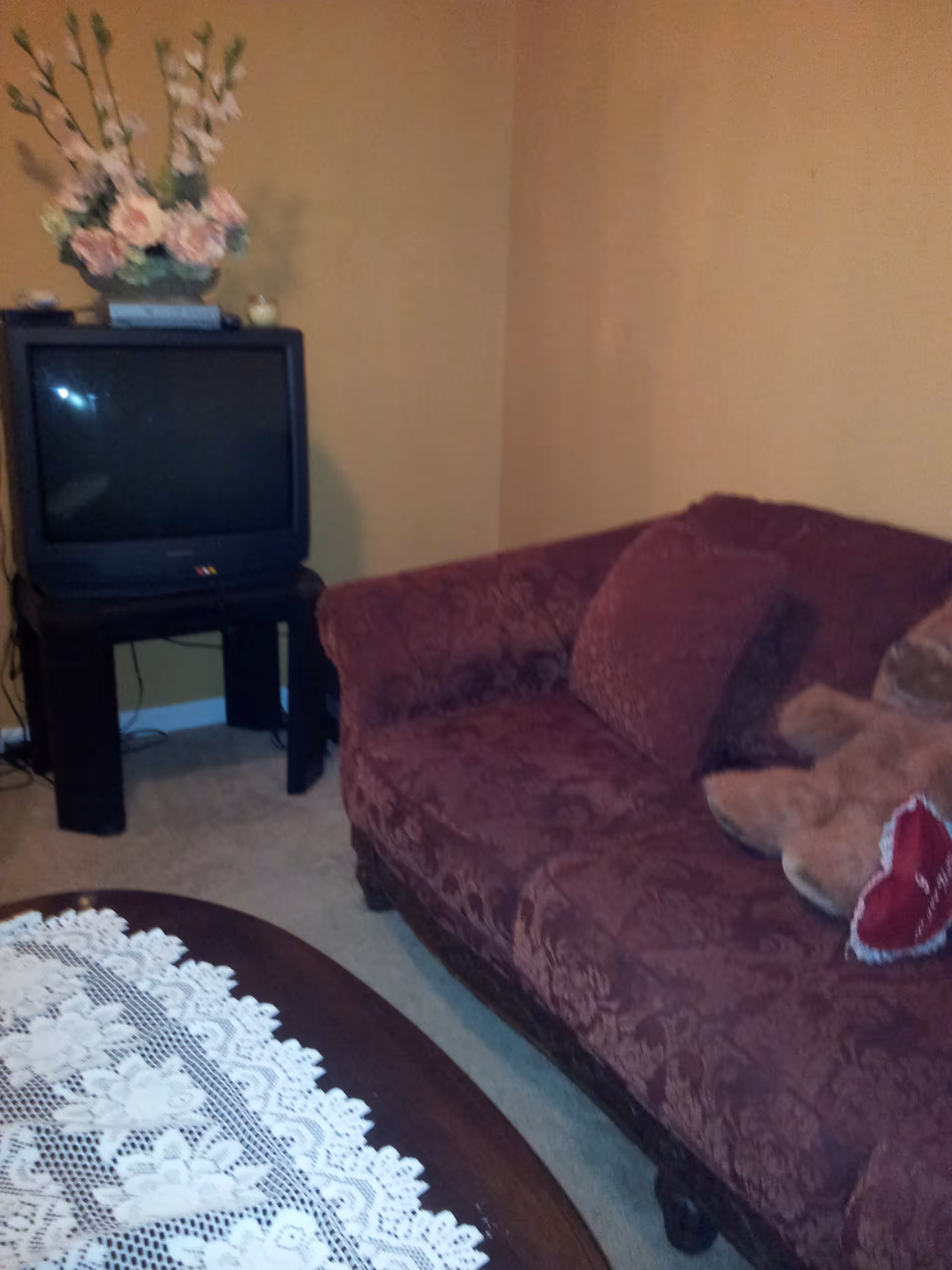 A cozy living room corner with a maroon patterned sofa adorned with cushions and a teddy bear. A wooden coffee table with a white lace tablecloth is partially visible in the foreground. In the corner, there is an old-style CRT television on a black stand with a floral arrangement on top. The walls are painted beige.