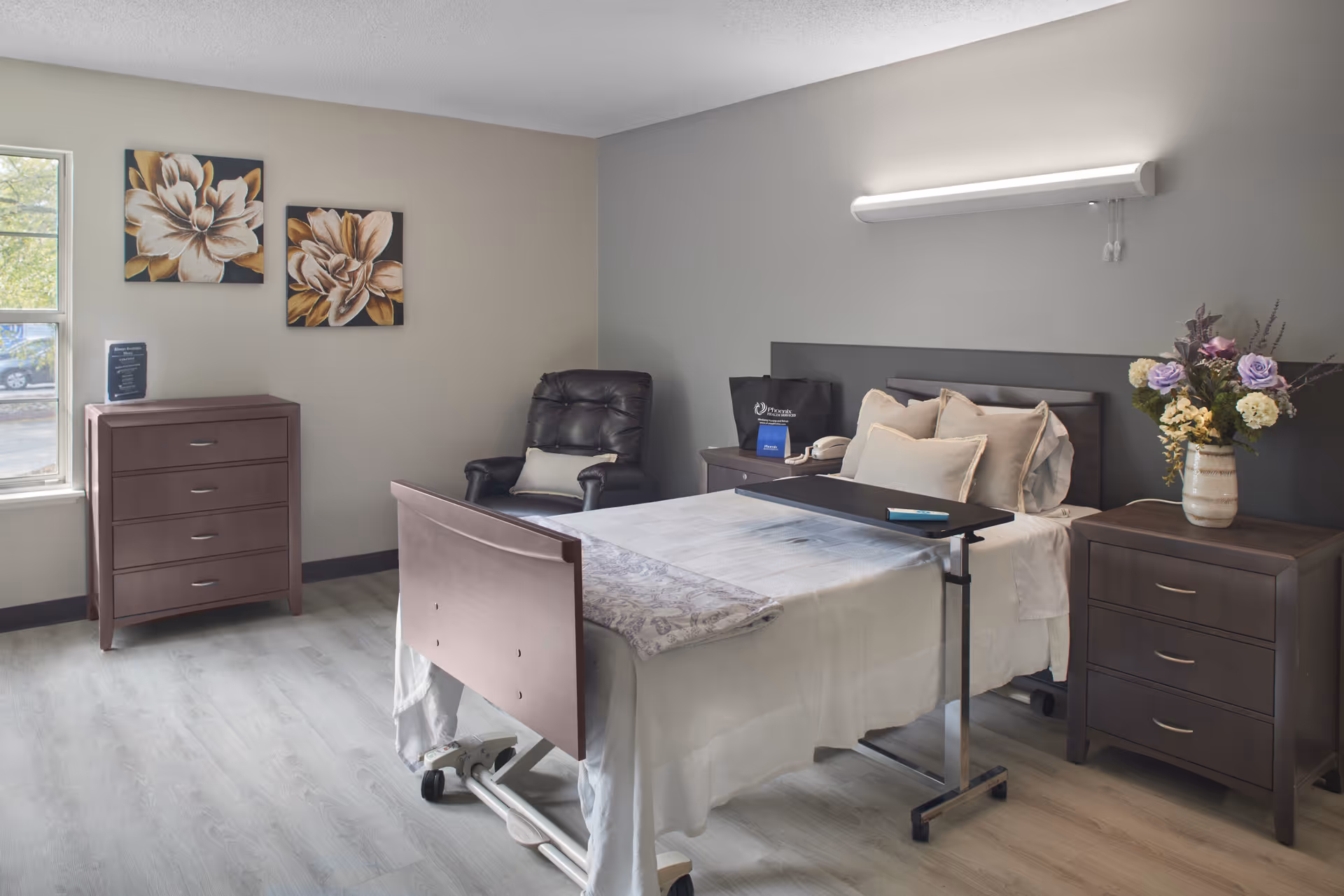 A furnished senior living bedroom with a hospital-style bed, overbed table, two bedside dressers, a recliner, and floral wall art and vase.
