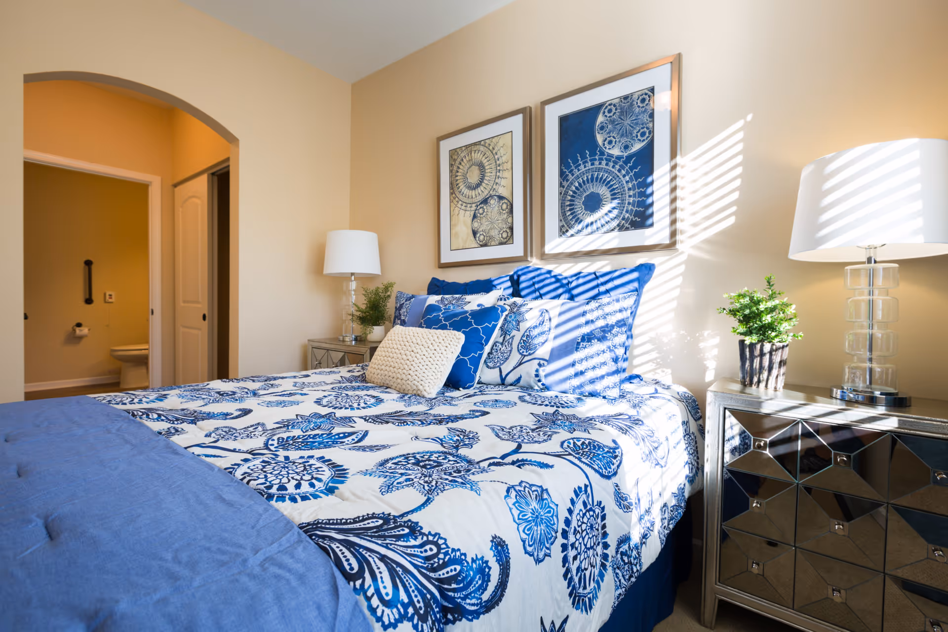 A bright bedroom with a bed covered in blue and white floral bedding, several pillows, two framed abstract art pieces on the wall, a mirrored nightstand with a lamp and a small plant, and an open doorway leading to a bathroom.