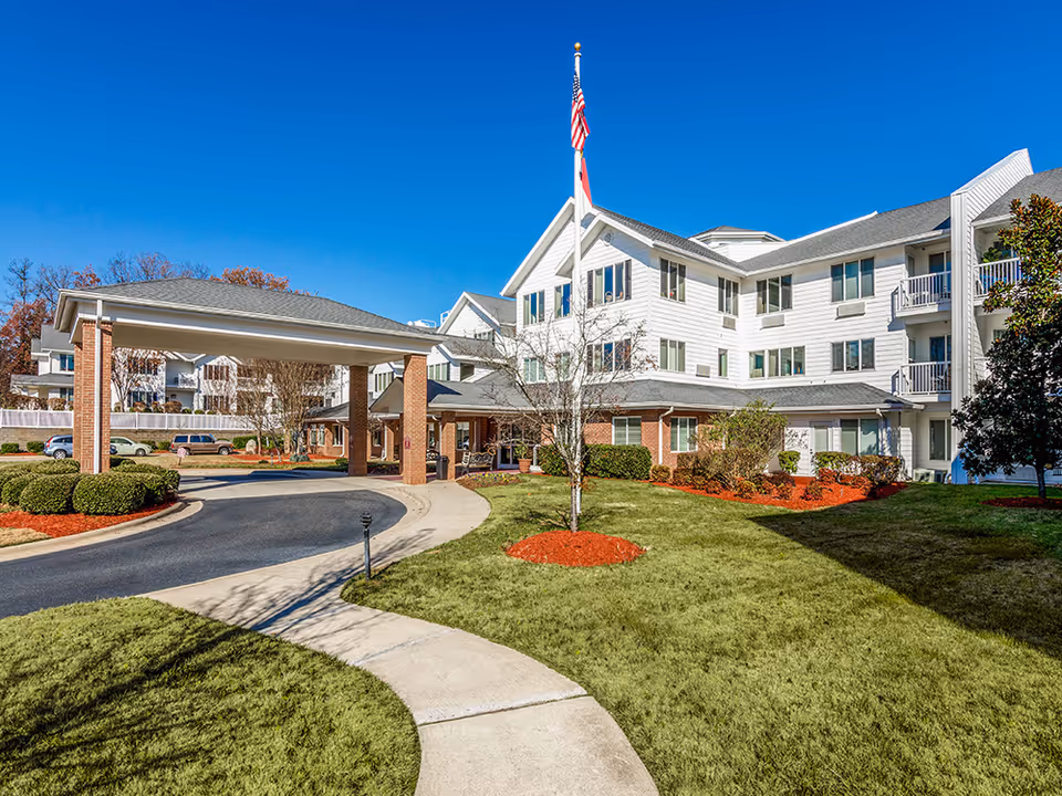 Exterior view of a senior living community building with white siding and brick accents. The building has multiple windows and balconies, a covered driveway entrance, a flagpole with an American flag, and well-maintained landscaping with green grass, bushes, and mulch beds under a clear blue sky.