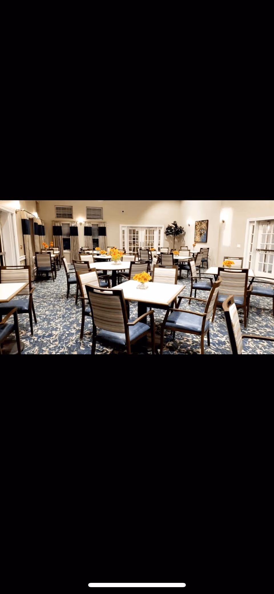 Spacious communal dining room with multiple square tables and chairs, floral centerpieces, patterned carpet, and bright lighting.