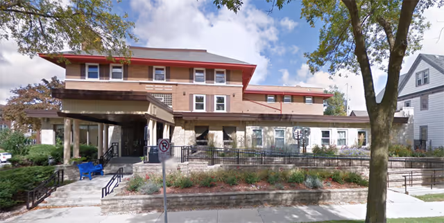 Exterior view of a multi-story senior living facility building with a covered entrance, a ramp, stairs, and landscaped garden beds in front. There is a large tree on the right side and a no parking sign near the entrance.