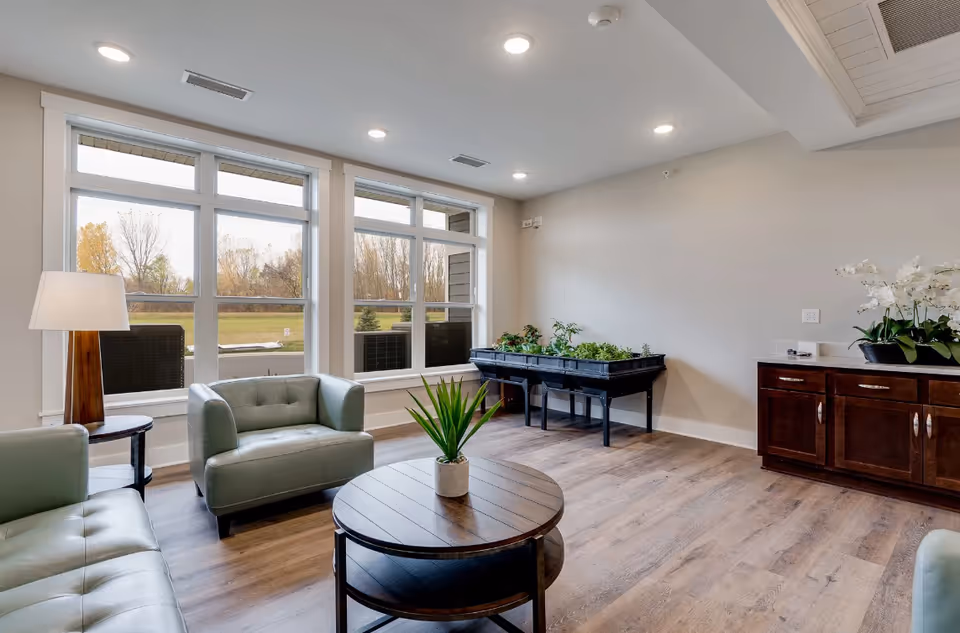 A bright and modern living room with large windows showing an outdoor view. The room features light green leather armchairs and a matching sofa, a round wooden coffee table with a small potted plant, a floor lamp, a wooden cabinet with white orchids, and a raised indoor garden planter with various green plants.