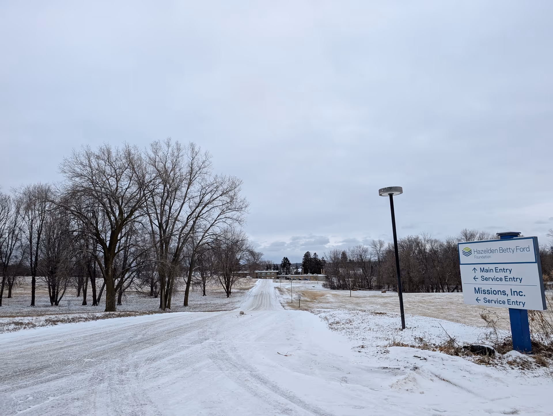 Snow-covered road leading to a building in the distance with leafless trees on both sides under a cloudy sky. A sign on the right side indicates directions for Hazelden Betty Ford Foundation and Missions, Inc. service entries.