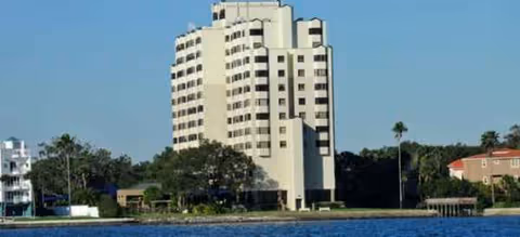 Tall beige senior living building by the water with trees along shoreline