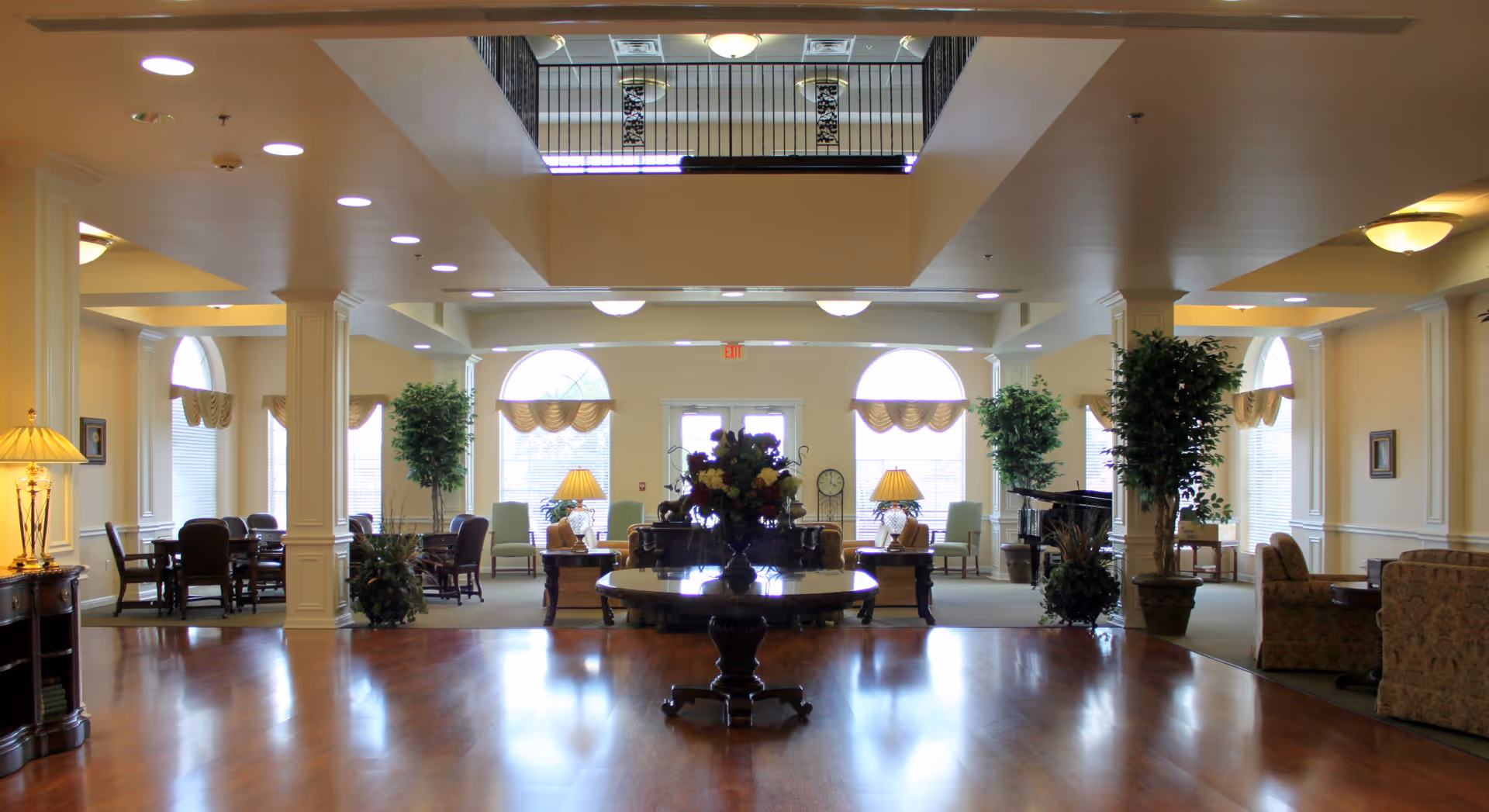 Spacious and well-lit common area in Oakview Assisted Living & Memory Care featuring polished wooden floors, a central round table with a floral arrangement, multiple seating areas with armchairs and tables, large arched windows with curtains, decorative lamps, potted plants, and a piano in the background.