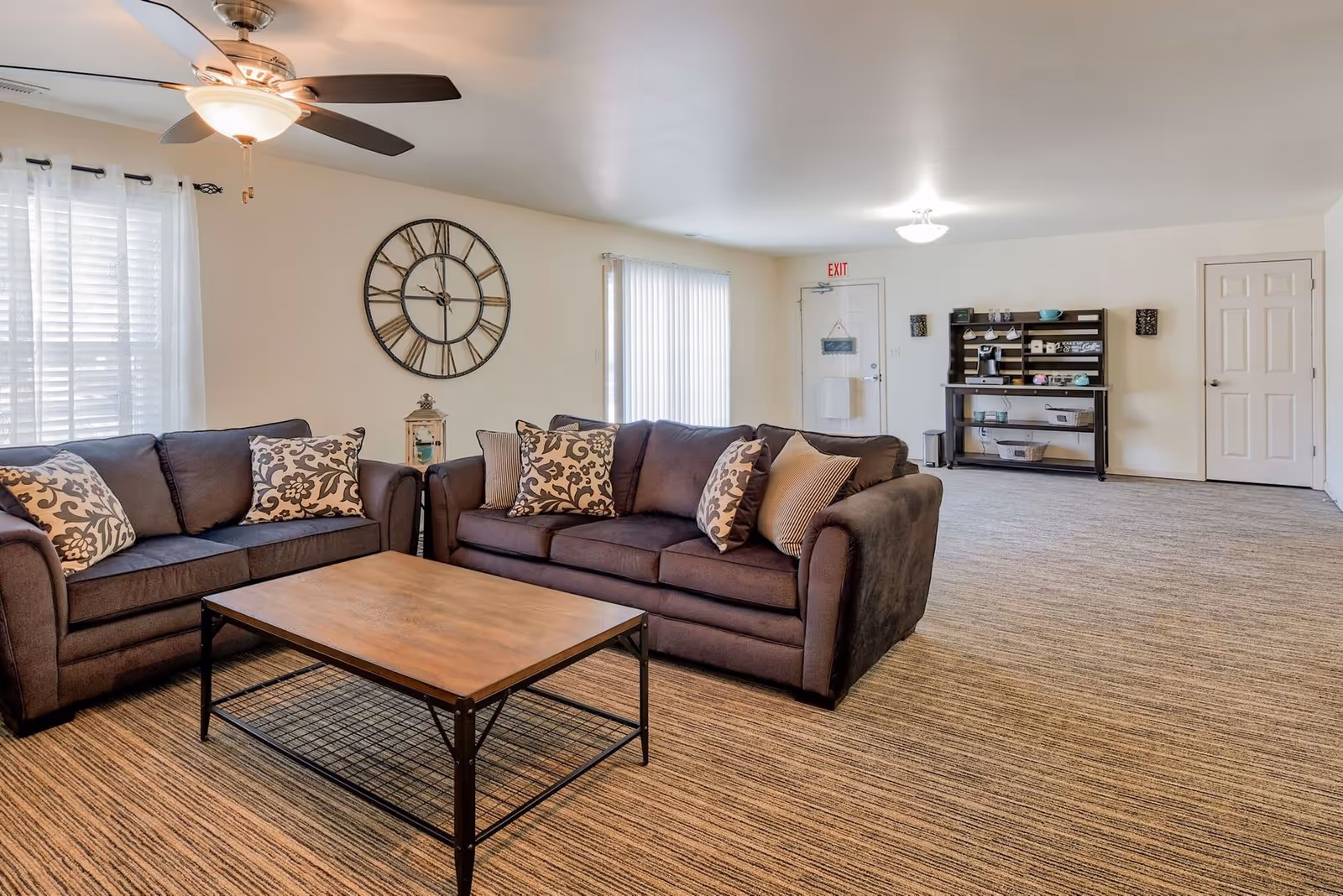 A spacious living room with two dark gray sofas adorned with patterned throw pillows, a wooden coffee table with a metal frame, a large wall clock, a ceiling fan with light, and a shelving unit with coffee mugs and a coffee maker near a white door with an exit sign above it.