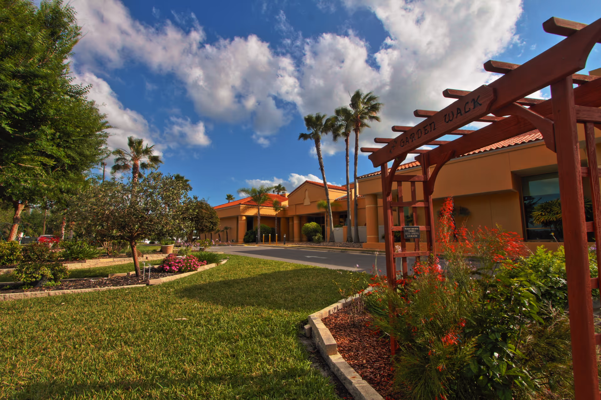 Exterior view of Cypress Lakes Village showing a landscaped garden area with green grass, flowering plants, palm trees, and a wooden pergola labeled 'Garden Walk' under a partly cloudy blue sky.