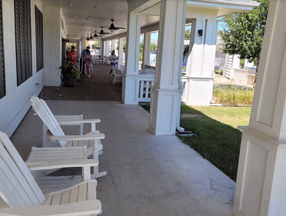 A covered outdoor patio area with white wooden chairs and tables. Several people, including a person using crutches, are walking or standing along the brick-paved section of the patio. The area is supported by white columns and overlooks a grassy lawn with trees and shrubs.