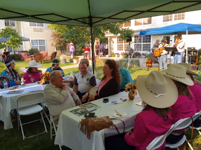 People seated at tables under a canopy in a grassy courtyard while musicians perform near the assisted living building.