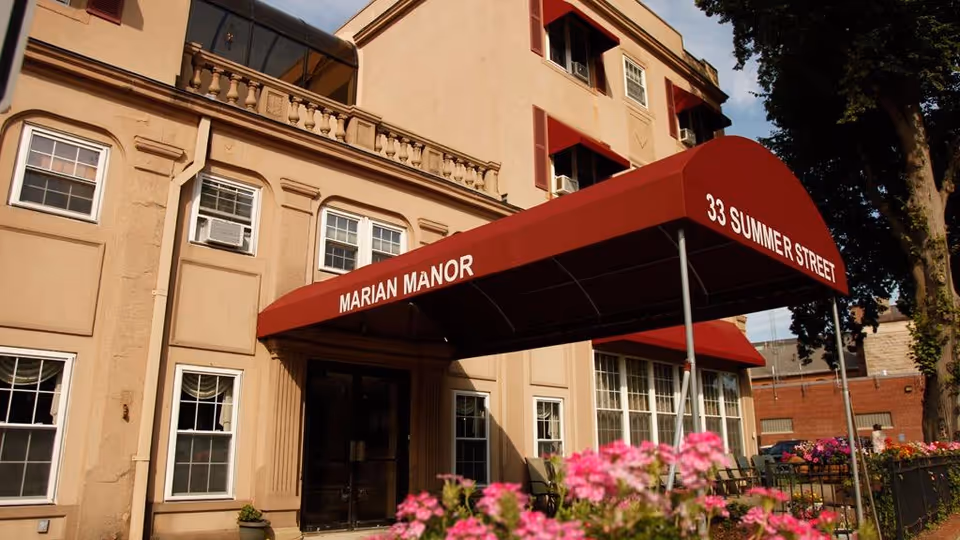 Exterior view of Marian Manor building with a beige facade and multiple windows. A red awning above the entrance displays the text 'MARIAN MANOR' and '33 SUMMER STREET'. Pink flowers and greenery are visible in the foreground.