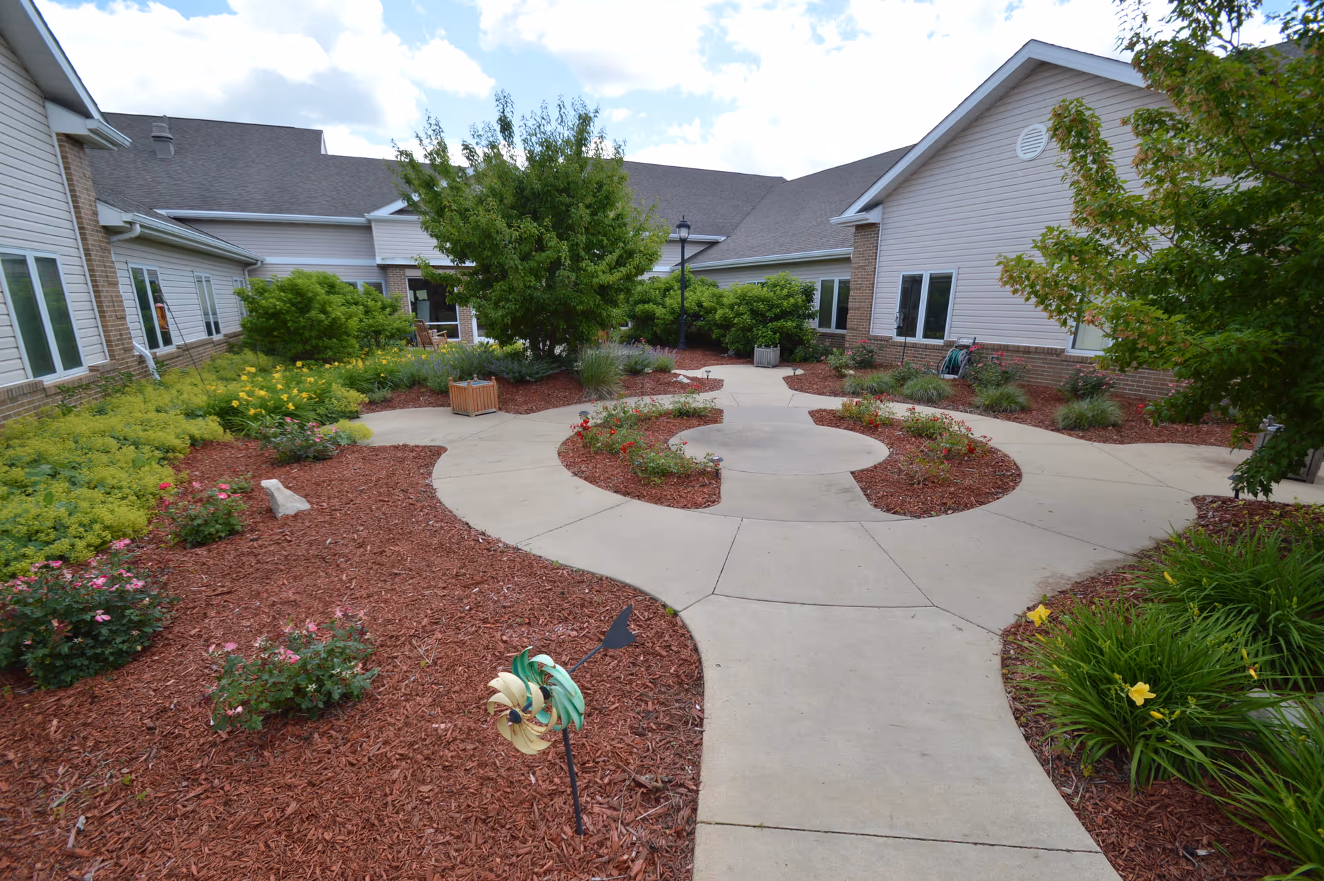Outdoor courtyard area at Southern Hills Specialty Care featuring a concrete walkway surrounded by landscaped garden beds with various shrubs, flowers, and mulch. The courtyard is enclosed by single-story building walls with multiple windows, and there is a tree and a lamppost in the center.