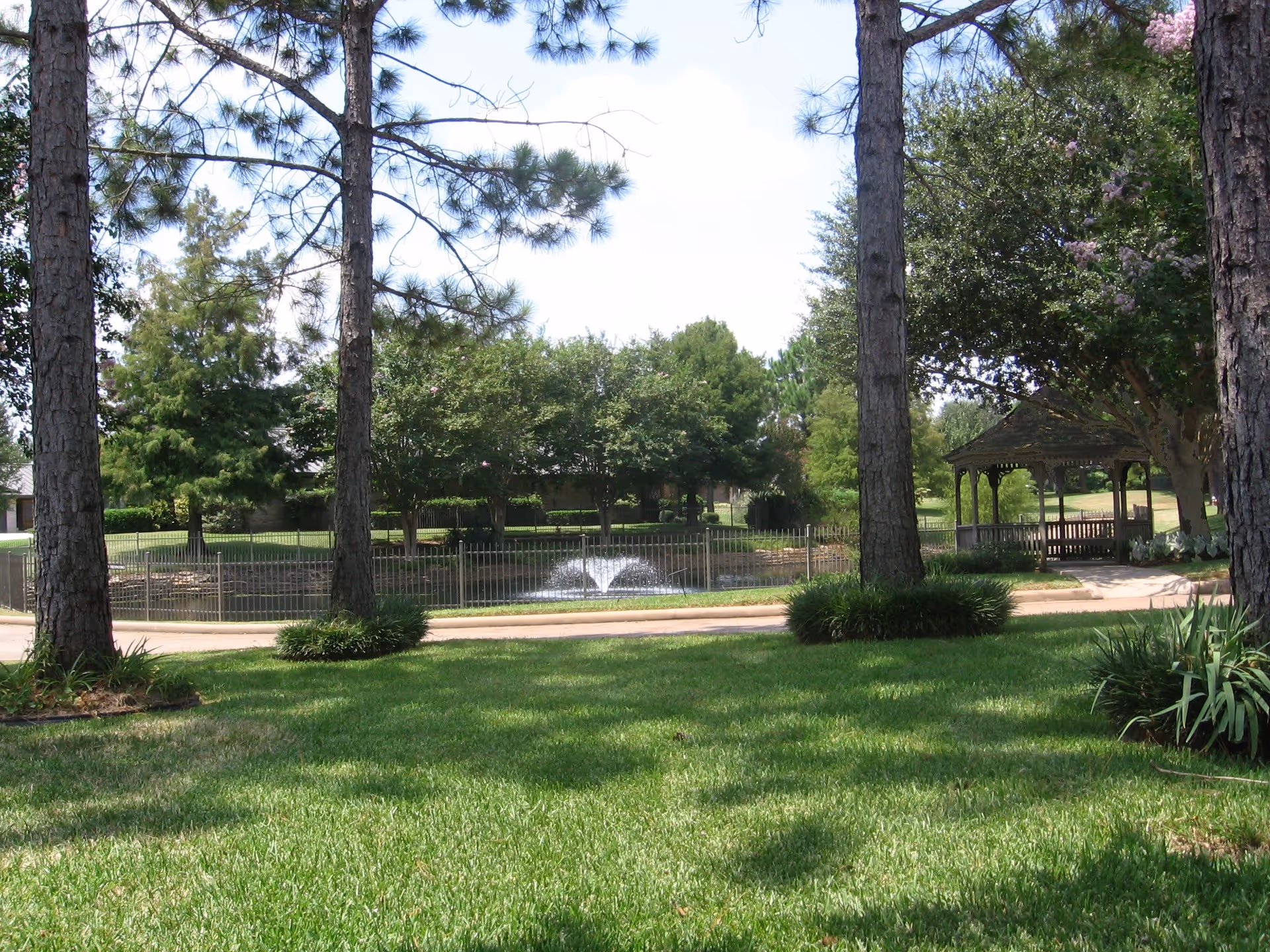 A peaceful outdoor scene at Kruse Village featuring a well-maintained grassy area with tall pine trees, a small pond with a water fountain, a paved walkway, and a wooden gazebo surrounded by lush greenery and flowering trees.