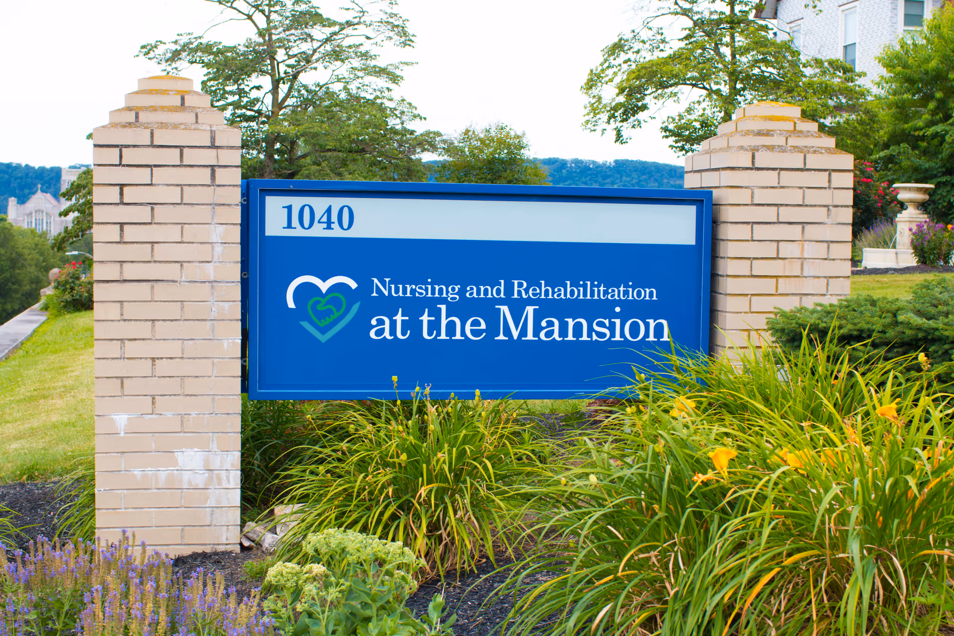 A blue sign with white text that reads 'Nursing and Rehabilitation at the Mansion' mounted between two beige brick pillars, surrounded by green plants and flowers with a building and trees in the background.
