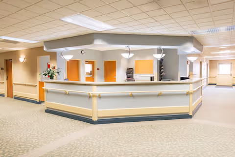 A bright and clean reception or nurse station area inside a healthcare facility with beige walls, carpeted floors, handrails along the walls, and several doors leading to other rooms. The ceiling has recessed lighting and hanging light fixtures above the counter.