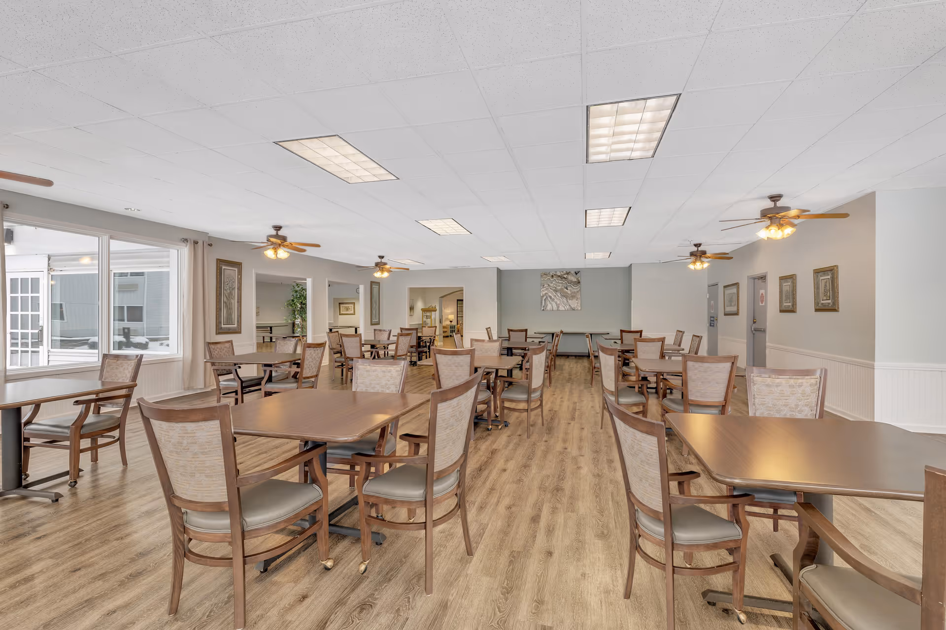 Spacious communal dining room with multiple wooden tables and chairs, ceiling fans, and large windows.