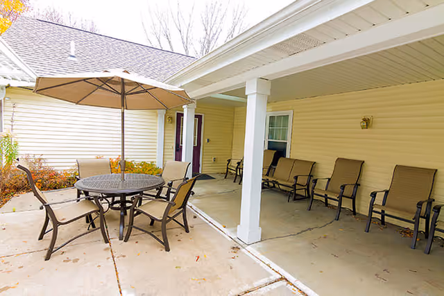Outdoor patio area with a round table and six chairs under a large umbrella. There are additional chairs lined up against the beige siding wall under a covered porch. Leafless trees are visible in the background.