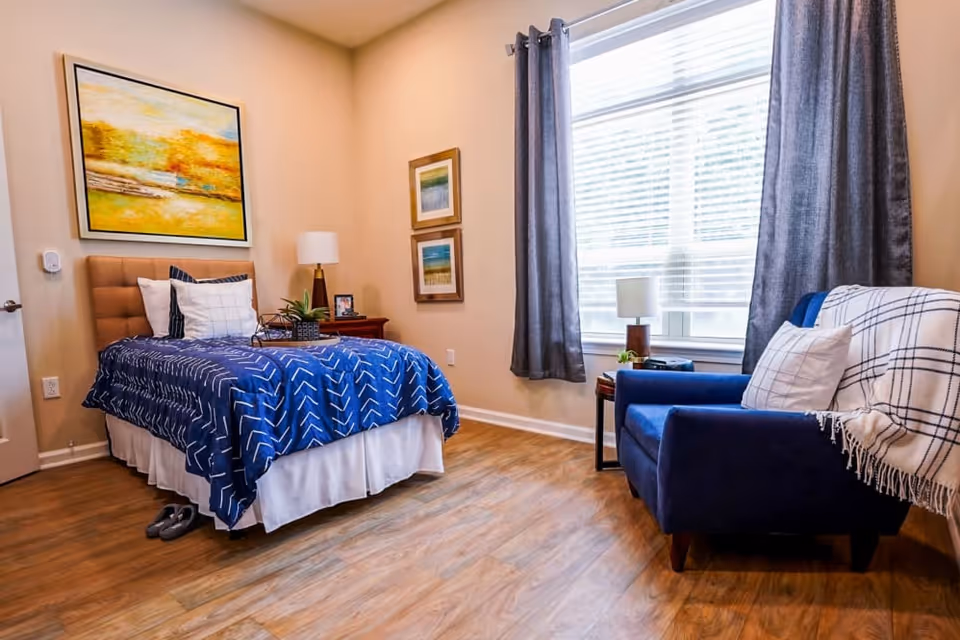 A cozy bedroom with a single bed covered in a blue patterned blanket and white pillows. Next to the bed is a wooden nightstand with a lamp, a small plant, and a framed photo. On the wall above the bed is a large colorful painting. Across from the bed is a blue armchair with a white and black checkered throw blanket and a white pillow. A window with gray curtains lets in natural light, illuminating the wooden floor.