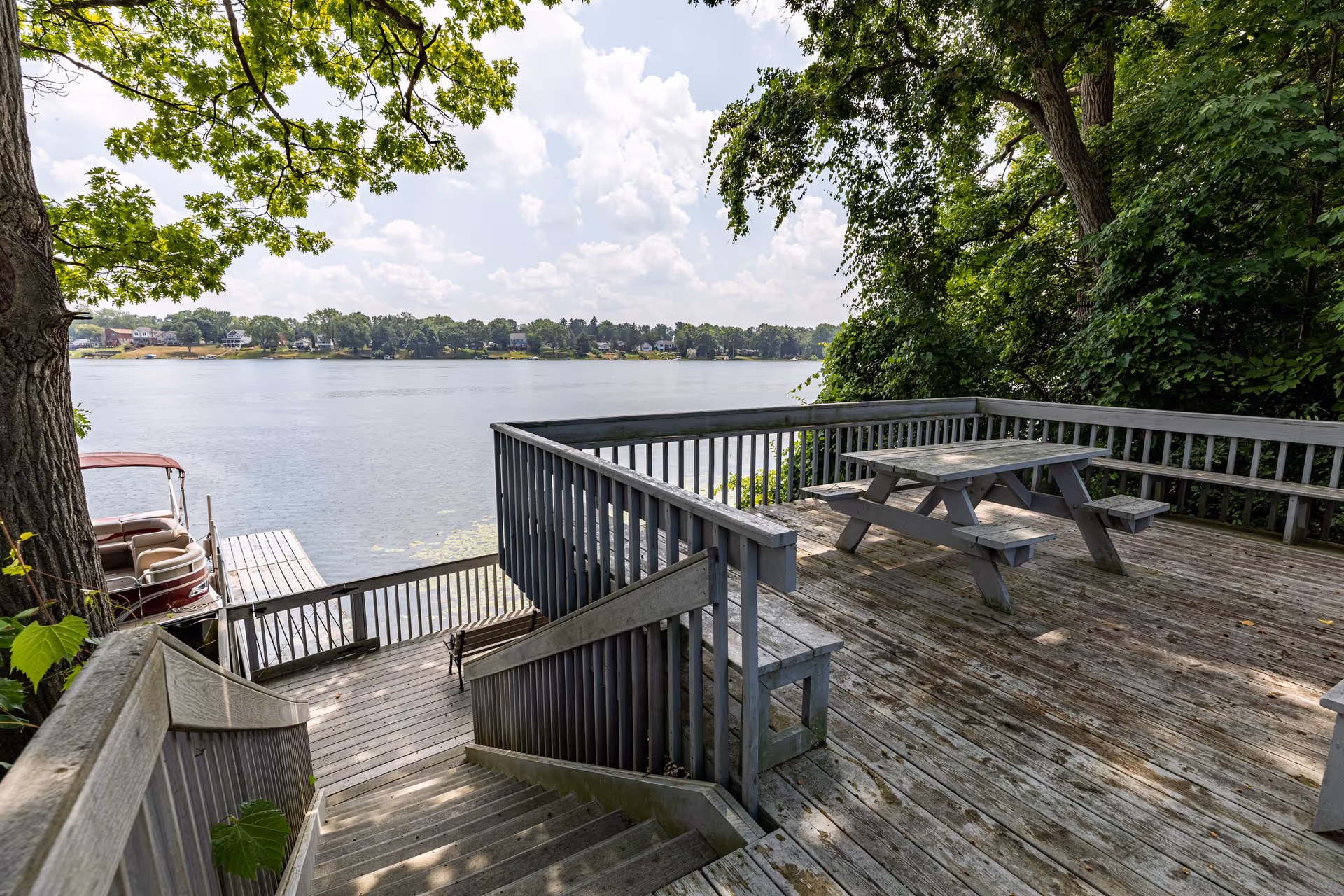 A wooden deck overlooking a serene lake, featuring a picnic table and surrounded by lush greenery. A boat is docked nearby, with a clear sky and distant houses visible across the water.