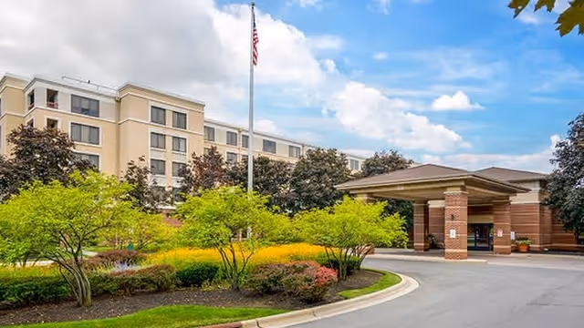 Front entrance of a multi-story senior living facility with a covered porte-cochère, landscaped gardens, and an American flag.