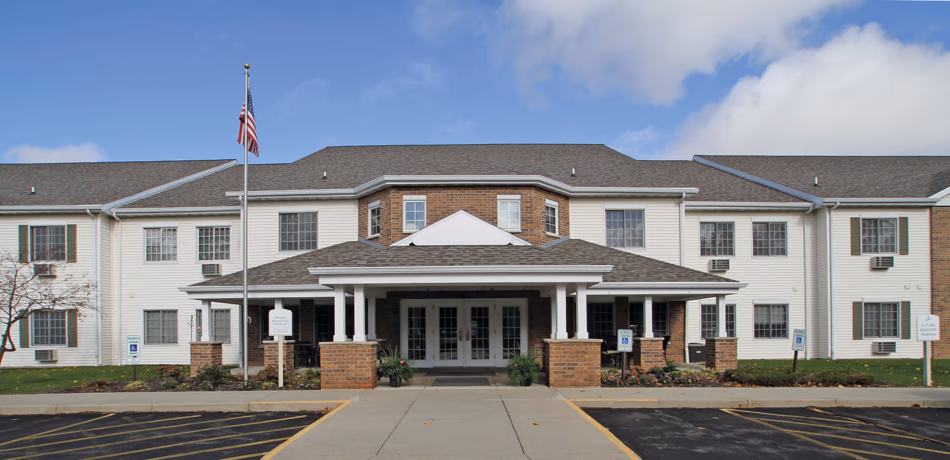 Front entrance of a two-story senior living building with a covered portico, brick accents, an American flag, and a parking area.