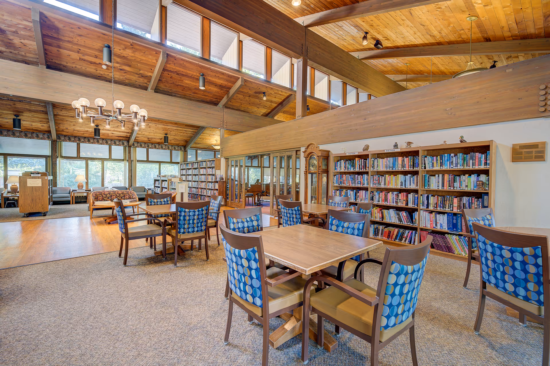 A spacious, well-lit community room with wooden vaulted ceilings and large windows near the roof. The room features several tables with chairs that have blue patterned backs, a bookshelf filled with books, a grandfather clock, and a cozy seating area with sofas and lamps near large windows. The room has a warm and inviting atmosphere with wood and carpet flooring.