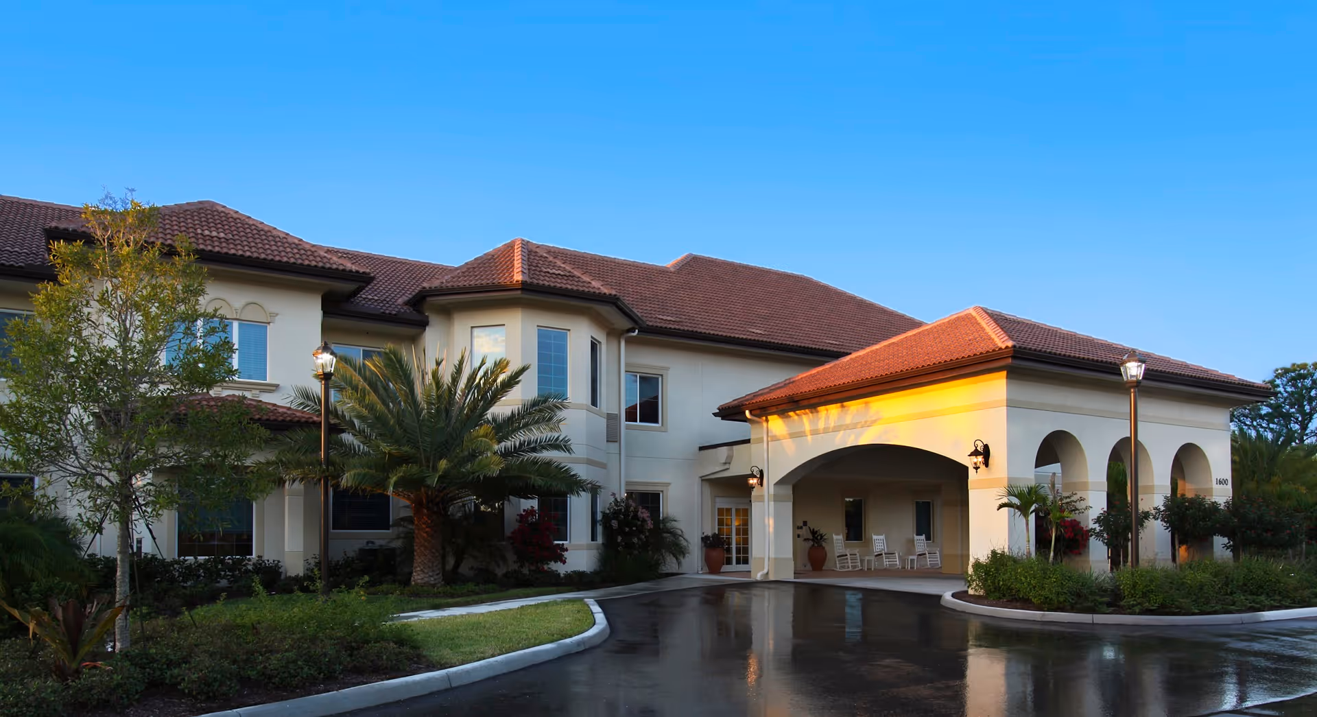 Front entrance of a two-story Mediterranean-style senior living building with a tiled roof, arched portico, palm trees, and a wet driveway.