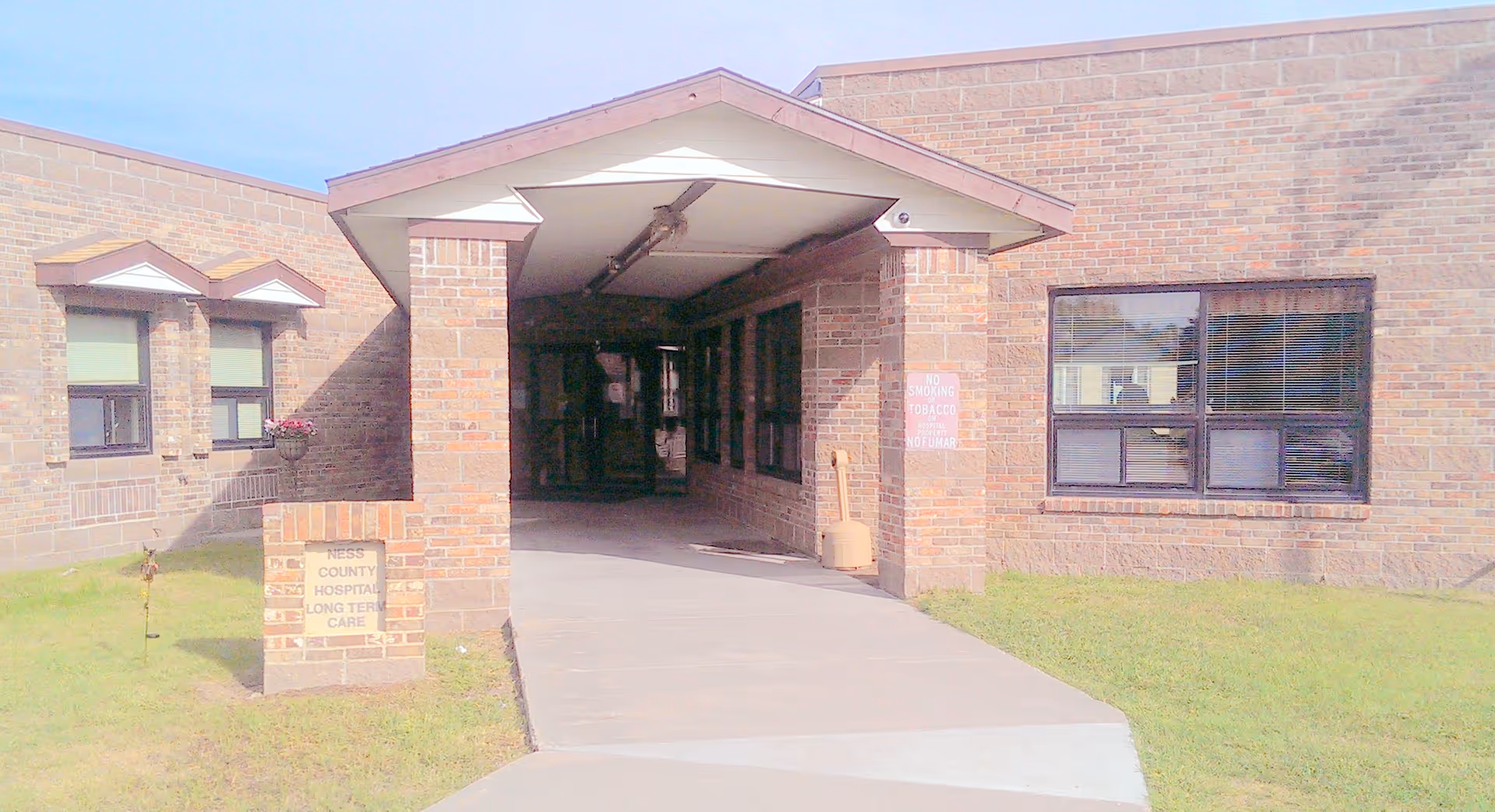 Entrance to Ness County Hospital Long Term Care building with a covered walkway, brick exterior walls, windows, and a small sign on the left side near the grass.